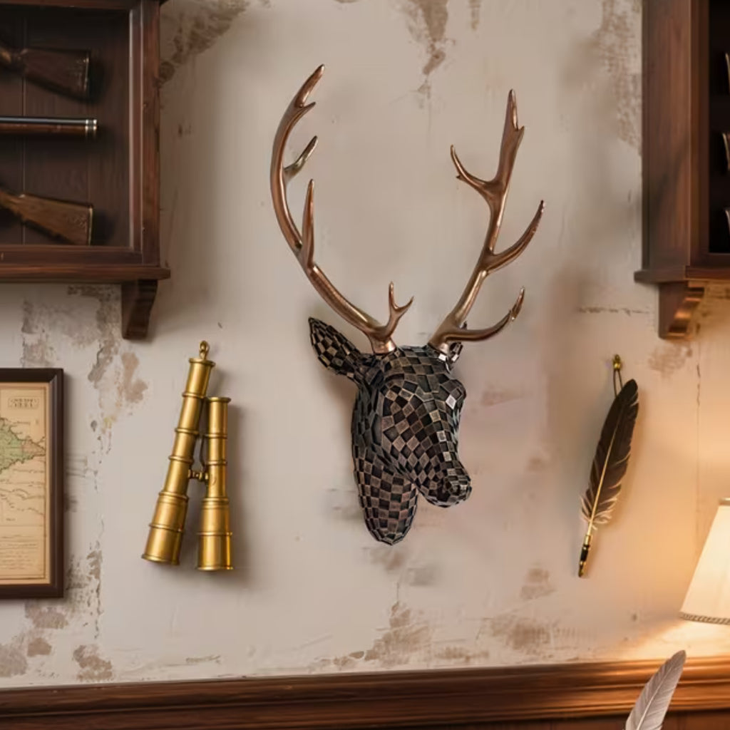 A decorative mounted deer head wall sculpture with large antlers, shown hanging on a distressed beige wall in a styled interior setting with brass binoculars, a feather, and wooden cabinetry.