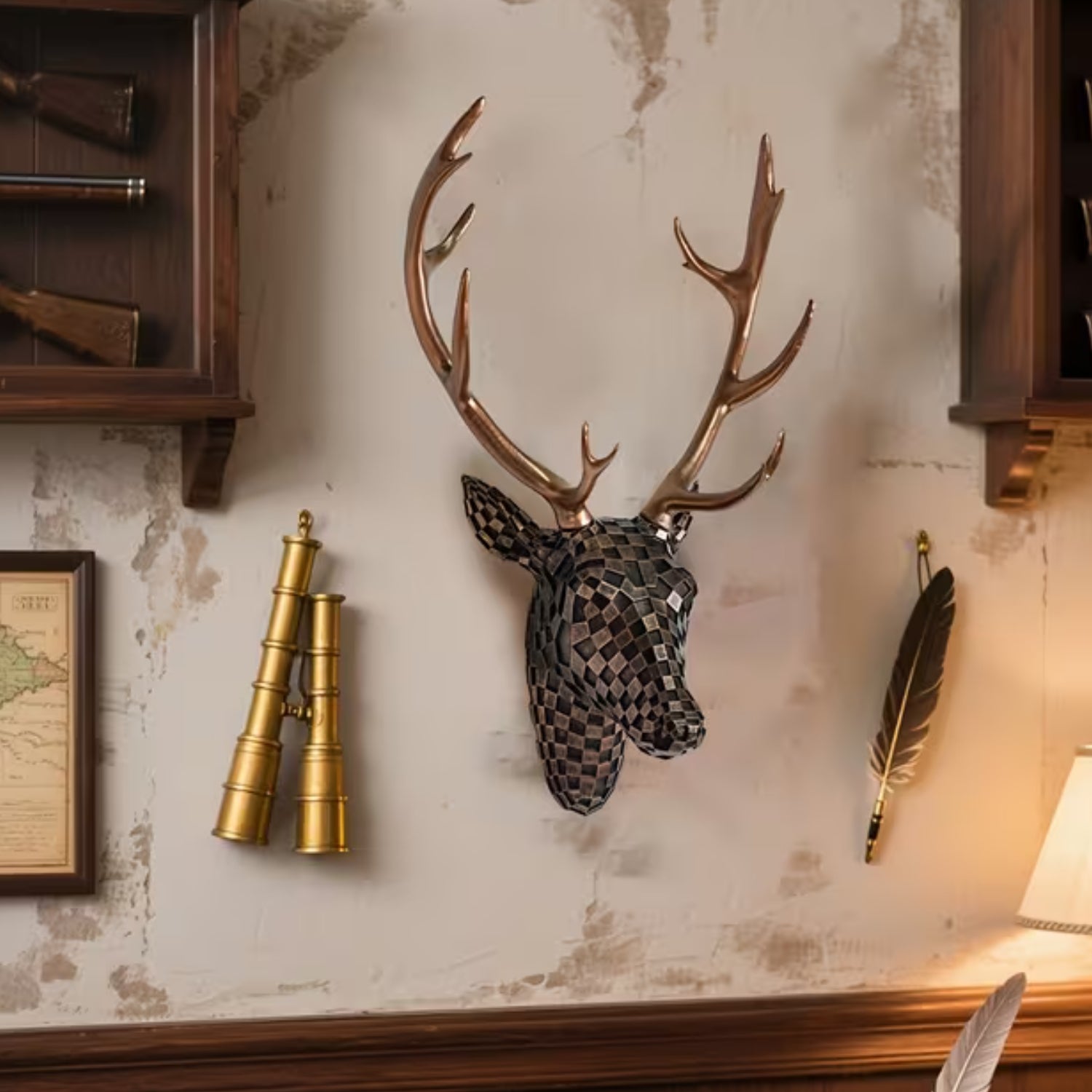 A decorative mounted deer head wall sculpture with large antlers, shown hanging on a distressed beige wall in a styled interior setting with brass binoculars, a feather, and wooden cabinetry.