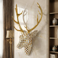 A decorative stag head wall mount with large gold antlers and a geometric checkered head in white and gold, displayed on a beige marble wall in an interior room setting.
