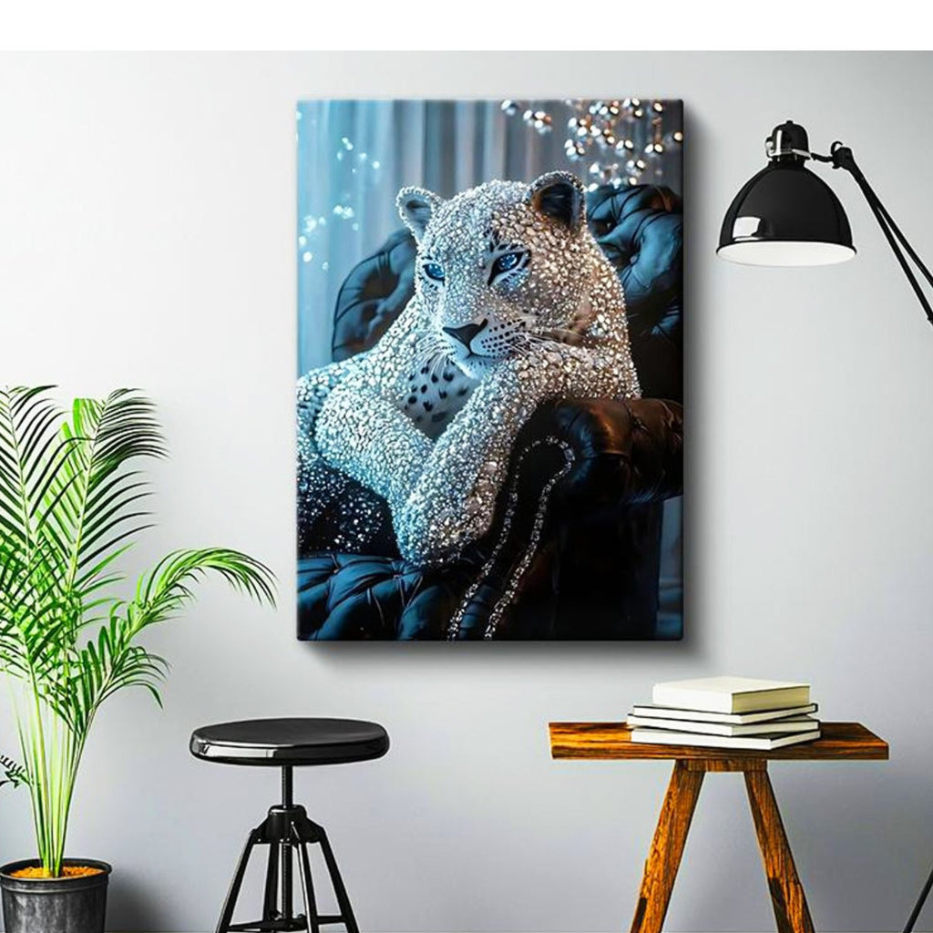 A rectangular wall art print showing a leopard reclining on a dark tufted chair, styled in a modern room with a black floor lamp, a potted plant, a stool, and a small wooden side table with books.