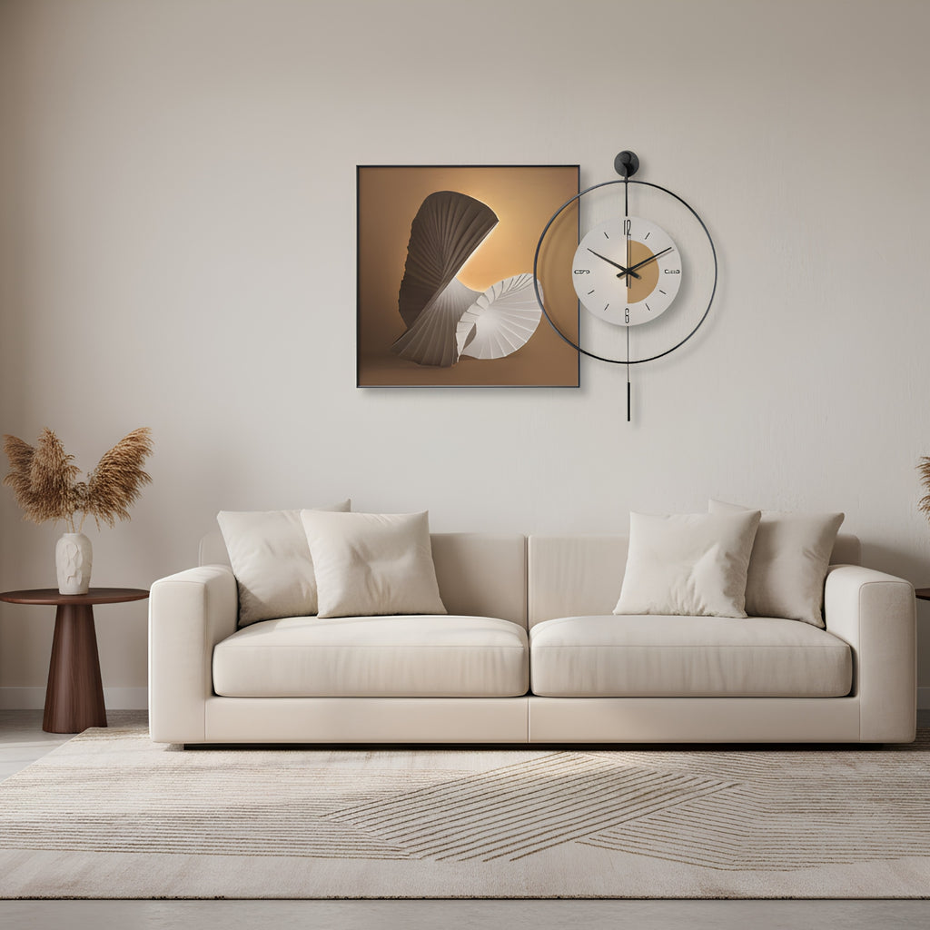 A modern living room scene featuring a cream-colored sofa, a small wooden side table with dried pampas grass, and a minimalist wall art piece depicting a fan and shell on a warm gradient background. A black metal ring clock is mounted on the wall above the sofa.