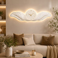 A decorative wall clock mounted above a beige sofa in a styled living room. The clock has a white sculptural leaf-like frame with warm LED lighting around it and a round clock face with gold hands and gold Roman numerals.
