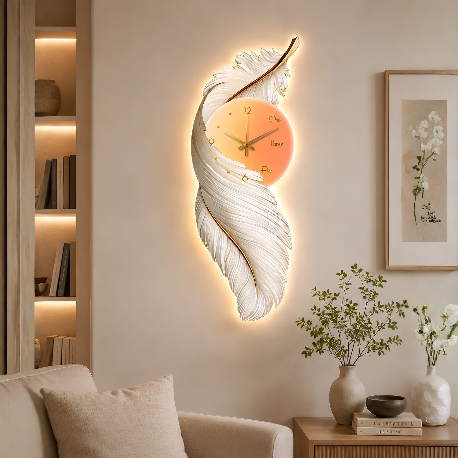 A decorative wall clock shaped like a glowing feather in a warm white and gold finish, mounted on a beige wall in a styled living room with a sofa, side table, framed art, and shelves.
