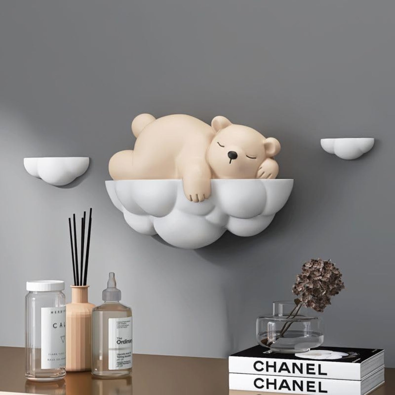 A beige sleeping bear wall shelf mounted on a gray wall above a tabletop, with a white cloud-shaped base. The scene includes small matching wall shelves, bottles, a reed diffuser, a glass vase with dried flowers, and a Chanel book on the table.