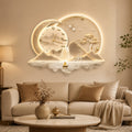 A decorative wall clock mounted above a beige sofa in a living room. The clock features a glowing circular design with a crescent moon, mountains, trees, a deer, and a small boat on the water, illuminated in warm light.