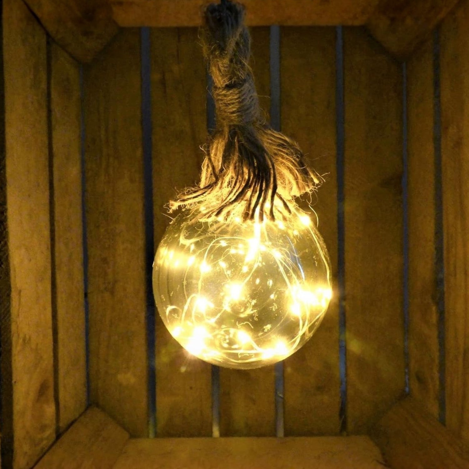 A clear glass light bulb filled with warm white fairy string lights, hanging from a rustic twine loop inside a wooden crate.