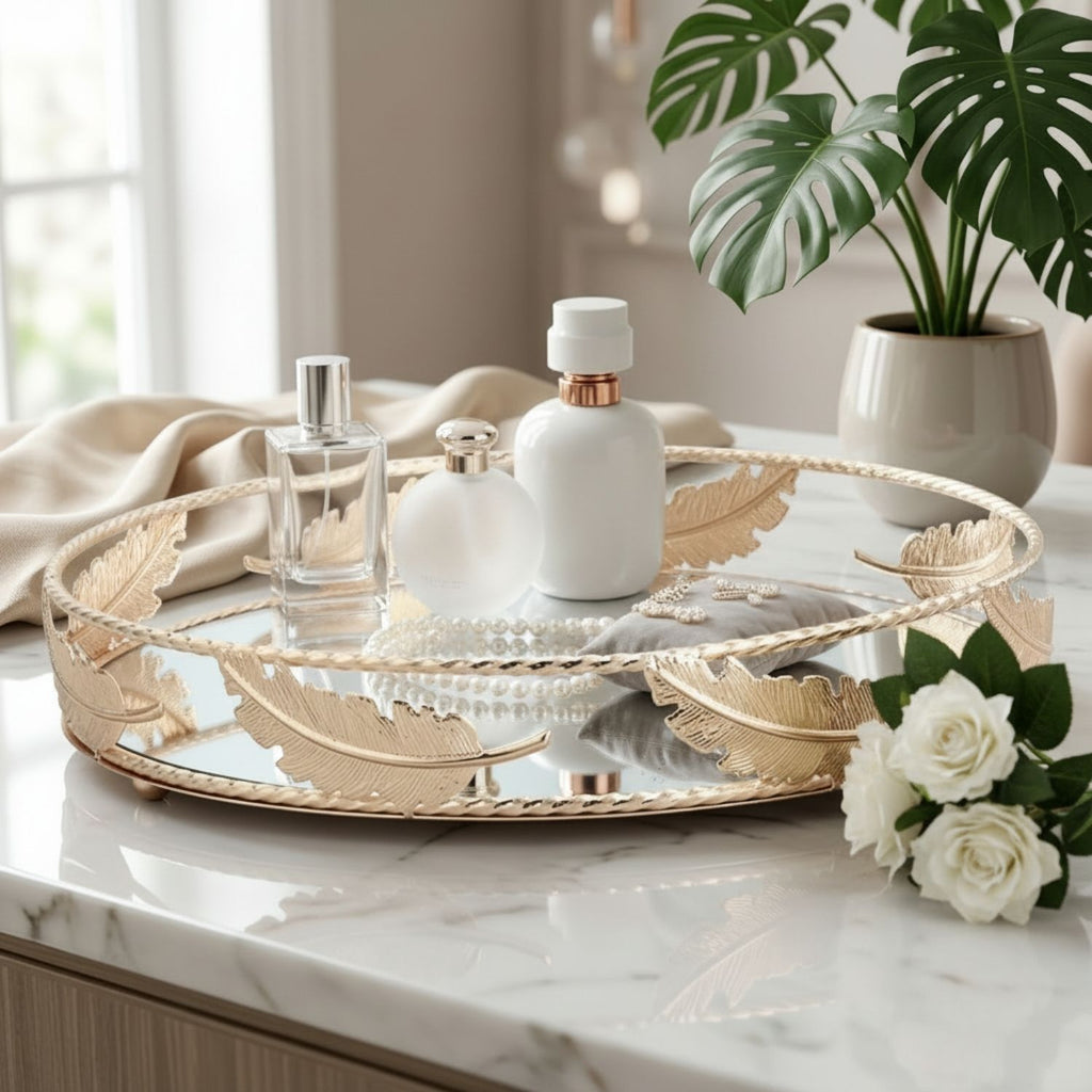 A round mirrored tray with gold-toned leaf accents and a rope trim, styled on a white marble countertop with perfume bottles, white flowers, a draped beige cloth, and potted plants in a bright indoor setting.