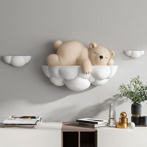 A beige sleeping bear figurine resting on a white cloud-shaped shelf mounted on a light gray wall above a console table with books, decor, and a plant.
