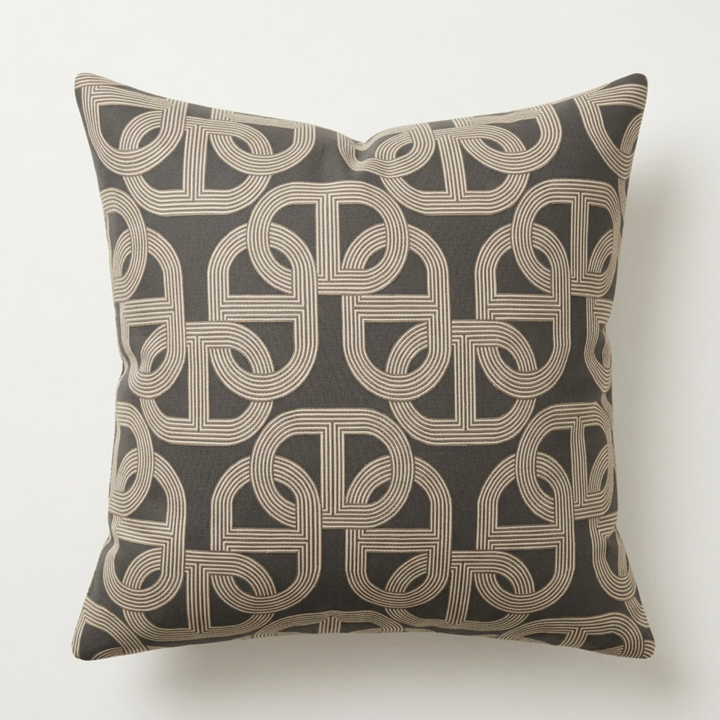 A square throw pillow featuring an intricate geometric interlocking line pattern in beige on a dark charcoal gray background, shown against a plain off-white wall.