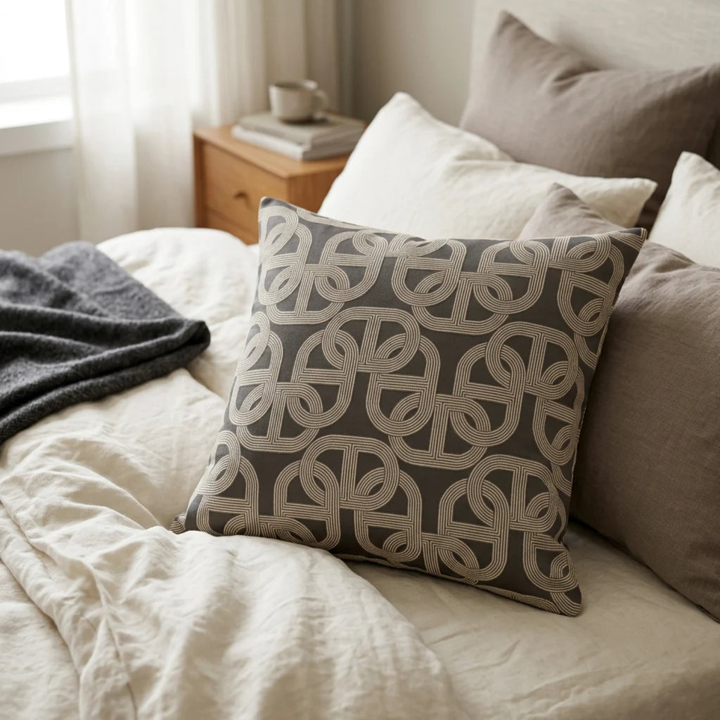 A square decorative throw pillow with a beige geometric interlocking pattern on a dark gray background, styled on a bed with neutral-colored bedding and pillows in a softly lit bedroom.