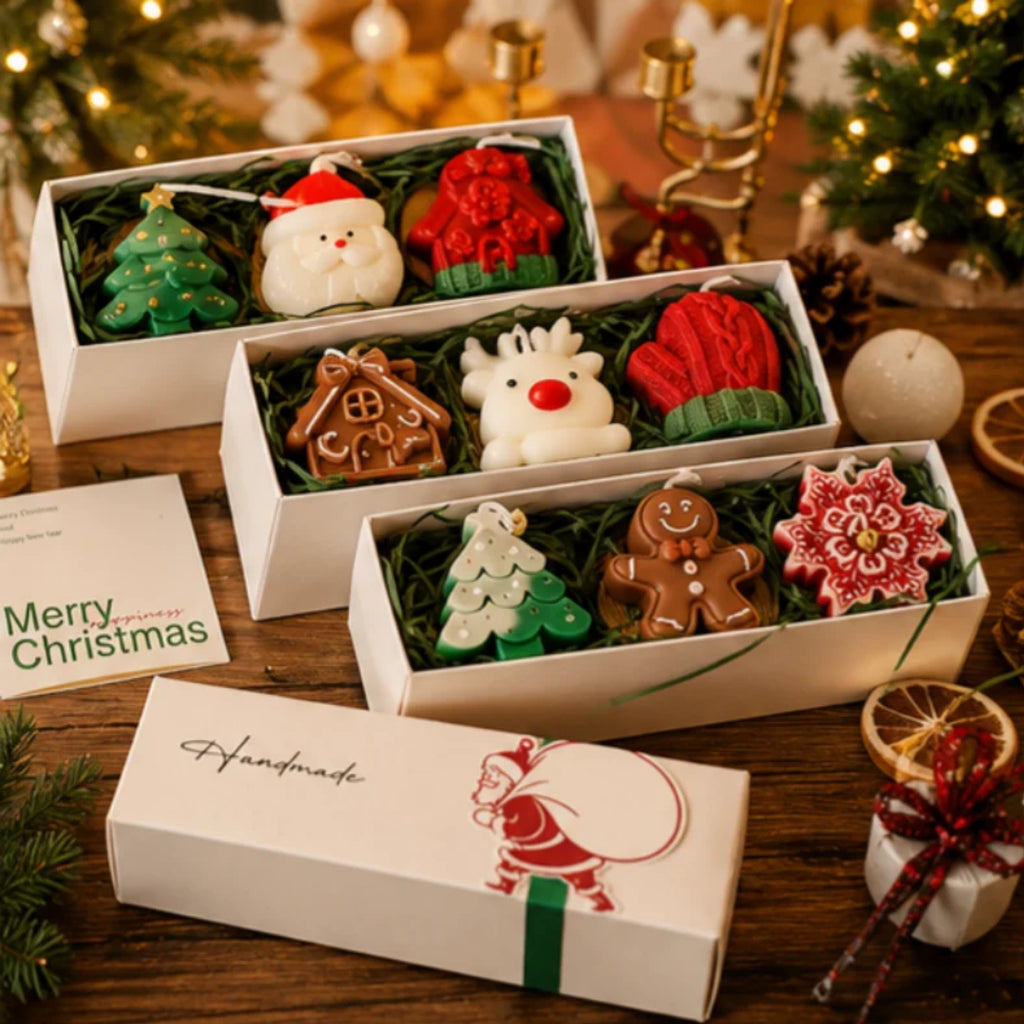 Three open white gift boxes filled with Christmas-themed shaped soaps, arranged on a wooden table with holiday decorations in the background. The soaps include a green tree, Santa, red hat, gingerbread house, white reindeer with red nose, red knit hat, gingerbread man, green tree, gingerbread cookie, and a red-and-white snowflake.