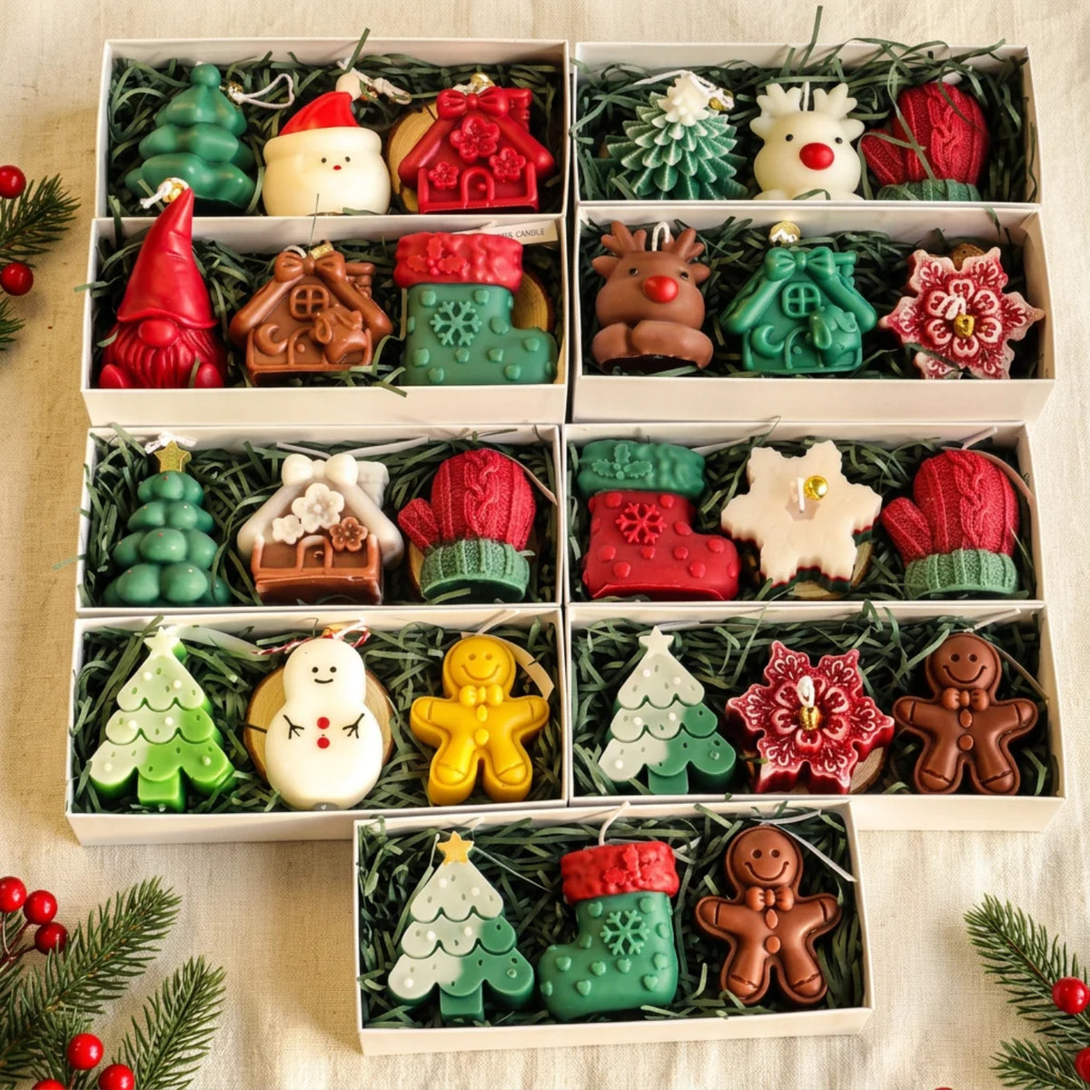 Six white gift boxes filled with Christmas-themed wax melts or soaps arranged in a grid, featuring shapes like trees, snowman, gingerbread, Santa, reindeer, mittens, boots, and floral ornaments, set on a beige fabric with pine branches and red berries.