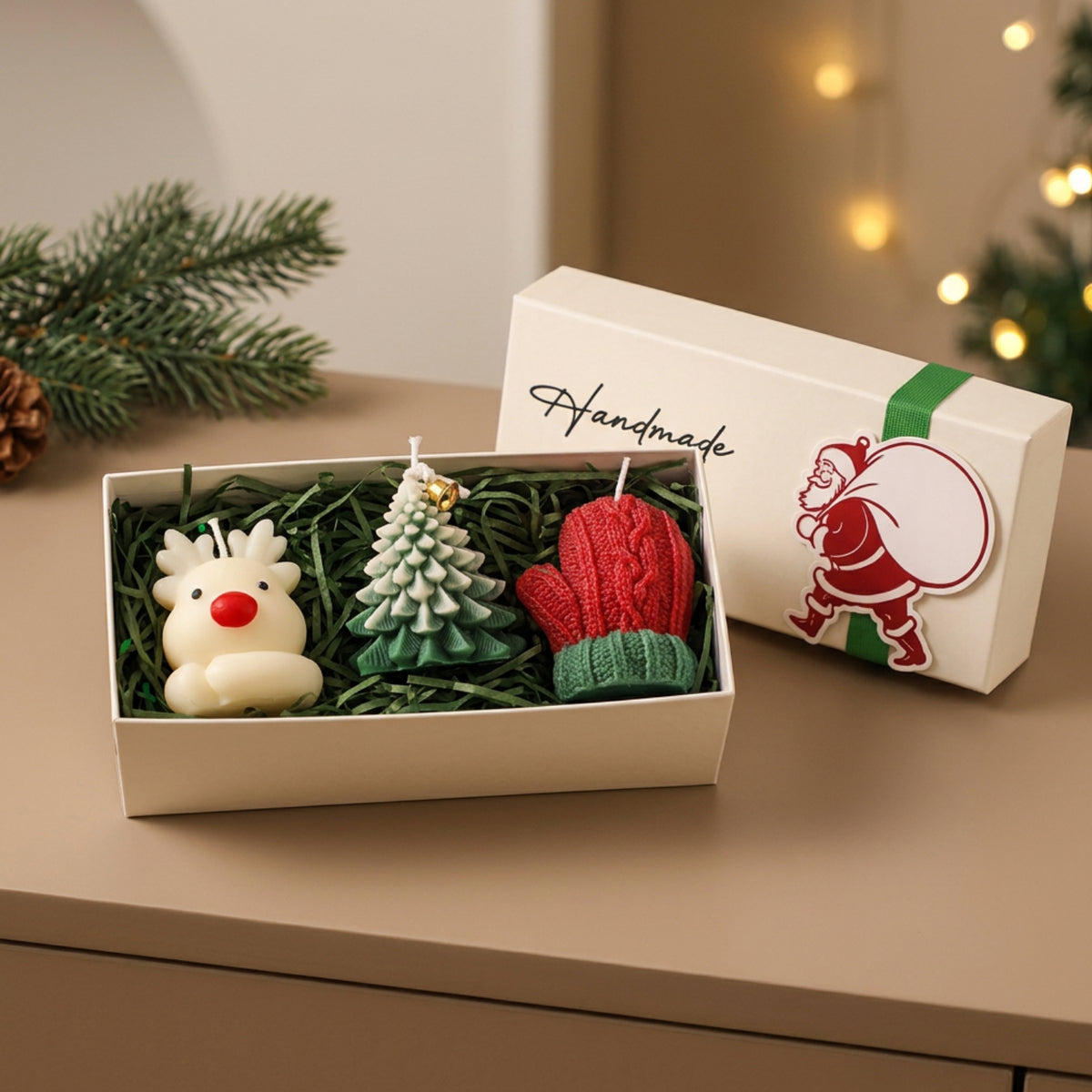 A boxed set of three Christmas-themed candles displayed on a tabletop, with the open box showing a white reindeer candle, a green tree candle, and a red knit mitten candle, alongside the white gift box lid printed with 'Handmade' and a Santa illustration.