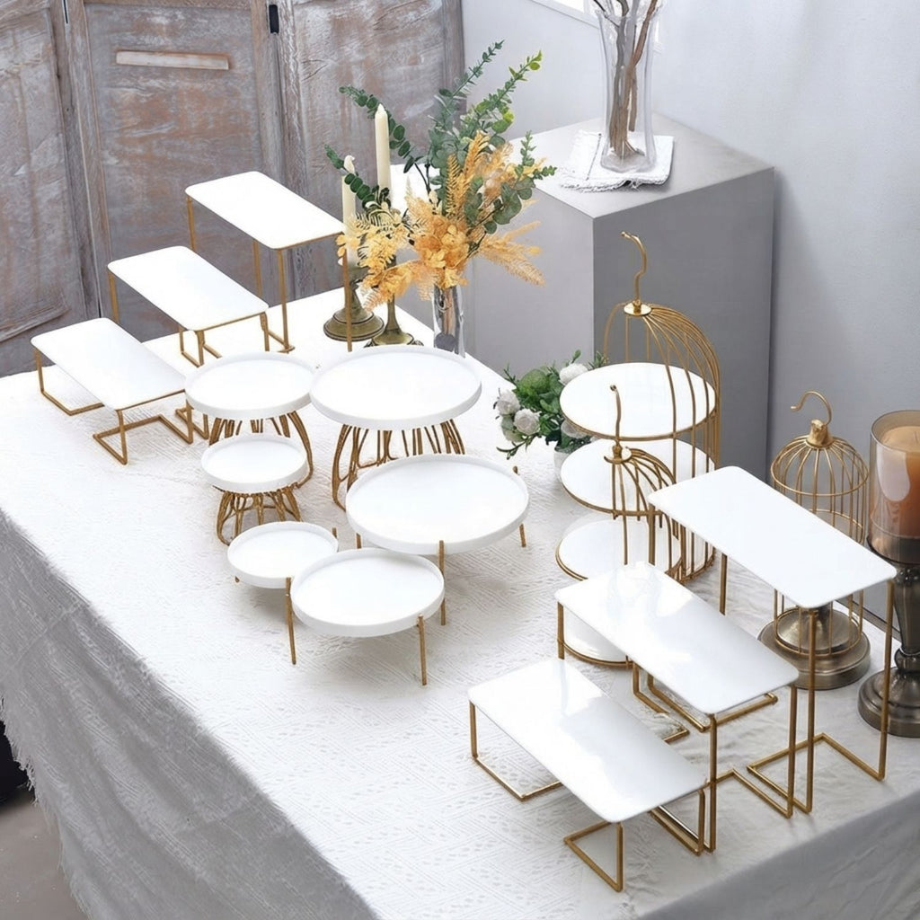 A set of white rectangular and round tiered display stands with gold metal frames, arranged on a white tablecloth in a styled interior setting.