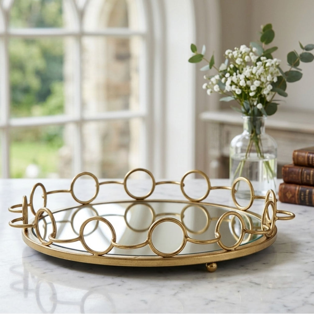 A round gold metal tray with a mirrored base and decorative interlocking circular wire design, styled on a marble table in a bright room with a vase of flowers and books in the background.