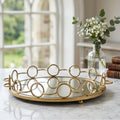 A round gold metal tray with a mirrored base and decorative interlocking circular wire design, styled on a marble table in a bright room with a vase of flowers and books in the background.