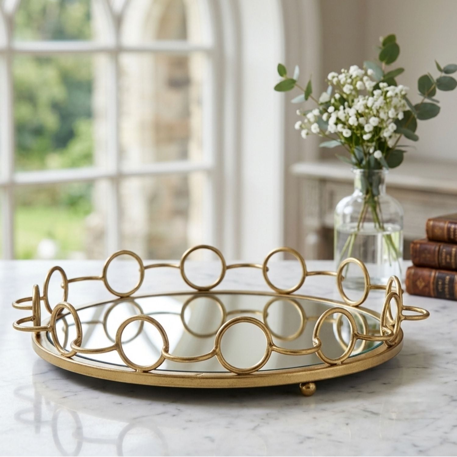 A round gold metal tray with a mirrored base and decorative interlocking circular wire design, styled on a marble table in a bright room with a vase of flowers and books in the background.