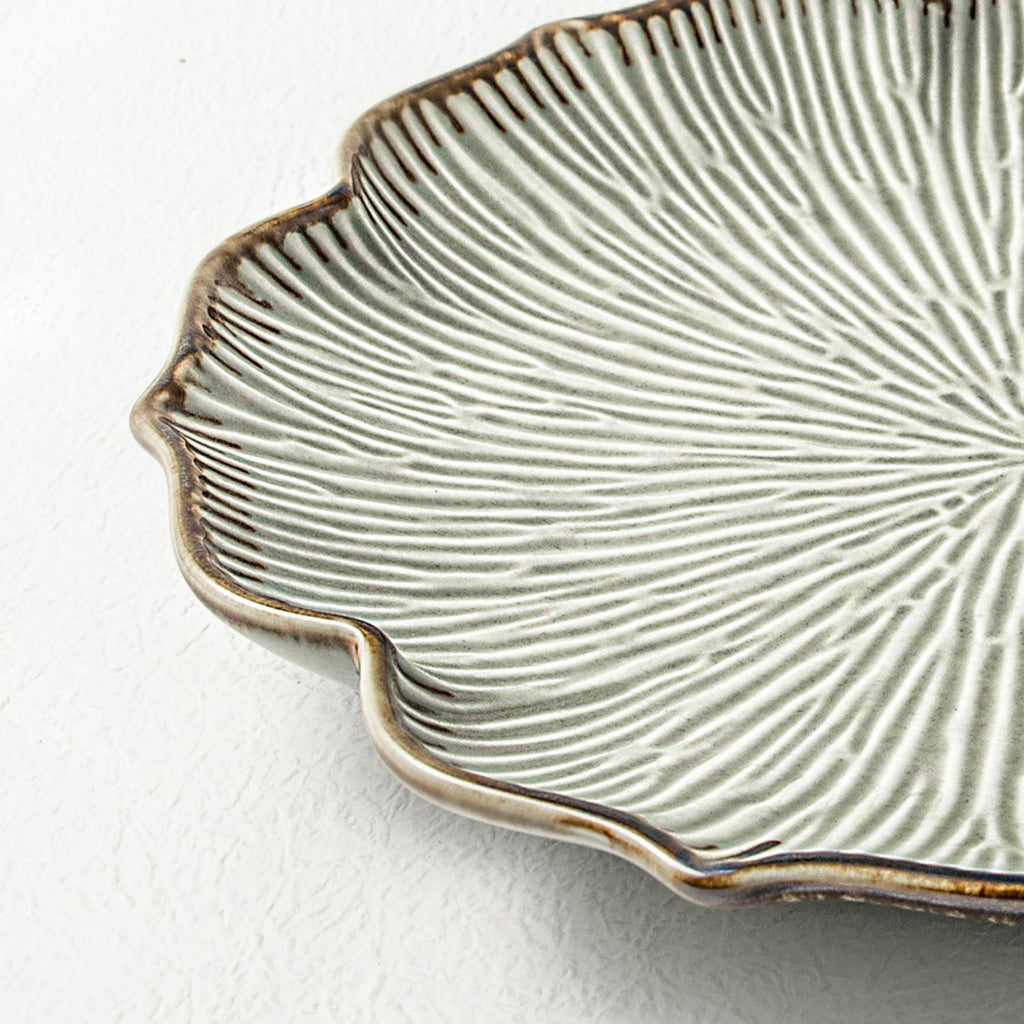 Close-up of a ceramic plate with a scalloped rim and a textured radial line pattern in warm beige and brown tones, set against a white textured background.