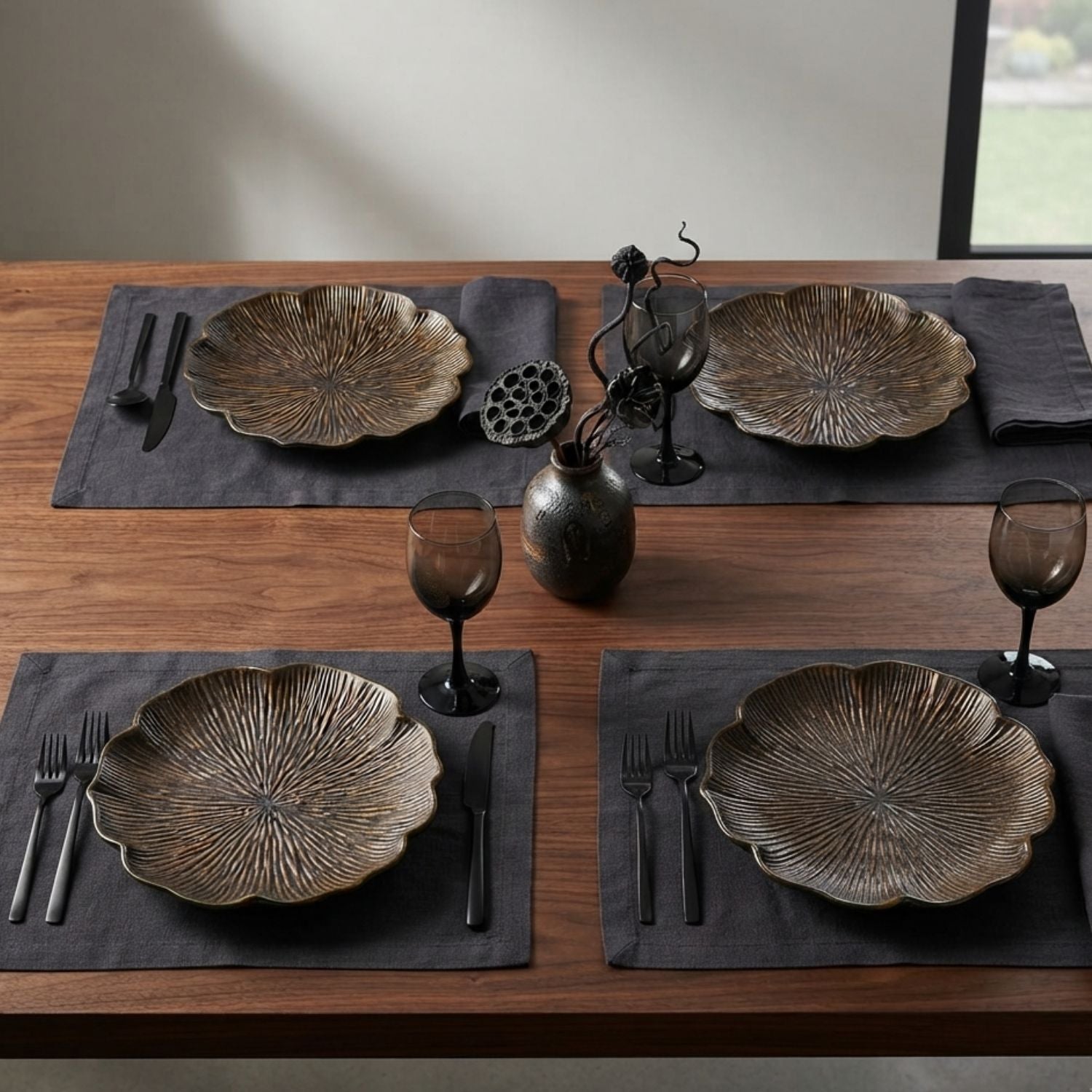 Four place settings on a wooden dining table, each featuring a textured bronze-toned plate, dark gray placemat, black cutlery, a dark glass goblet, and a small decorative vase in the center.