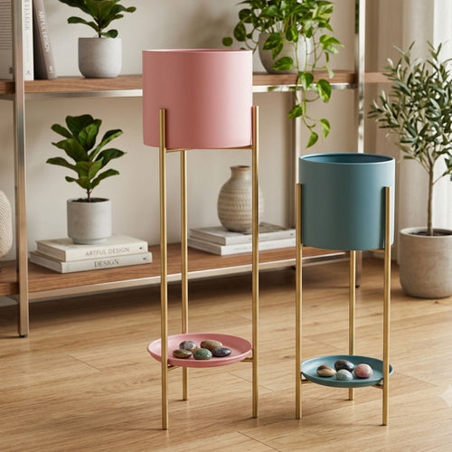 Two modern floor planters with cylindrical pots in pink and blue, supported by slim gold metal stands with a small matching tray at the bottom, styled in a warm, sunlit room with wooden floors and shelves.