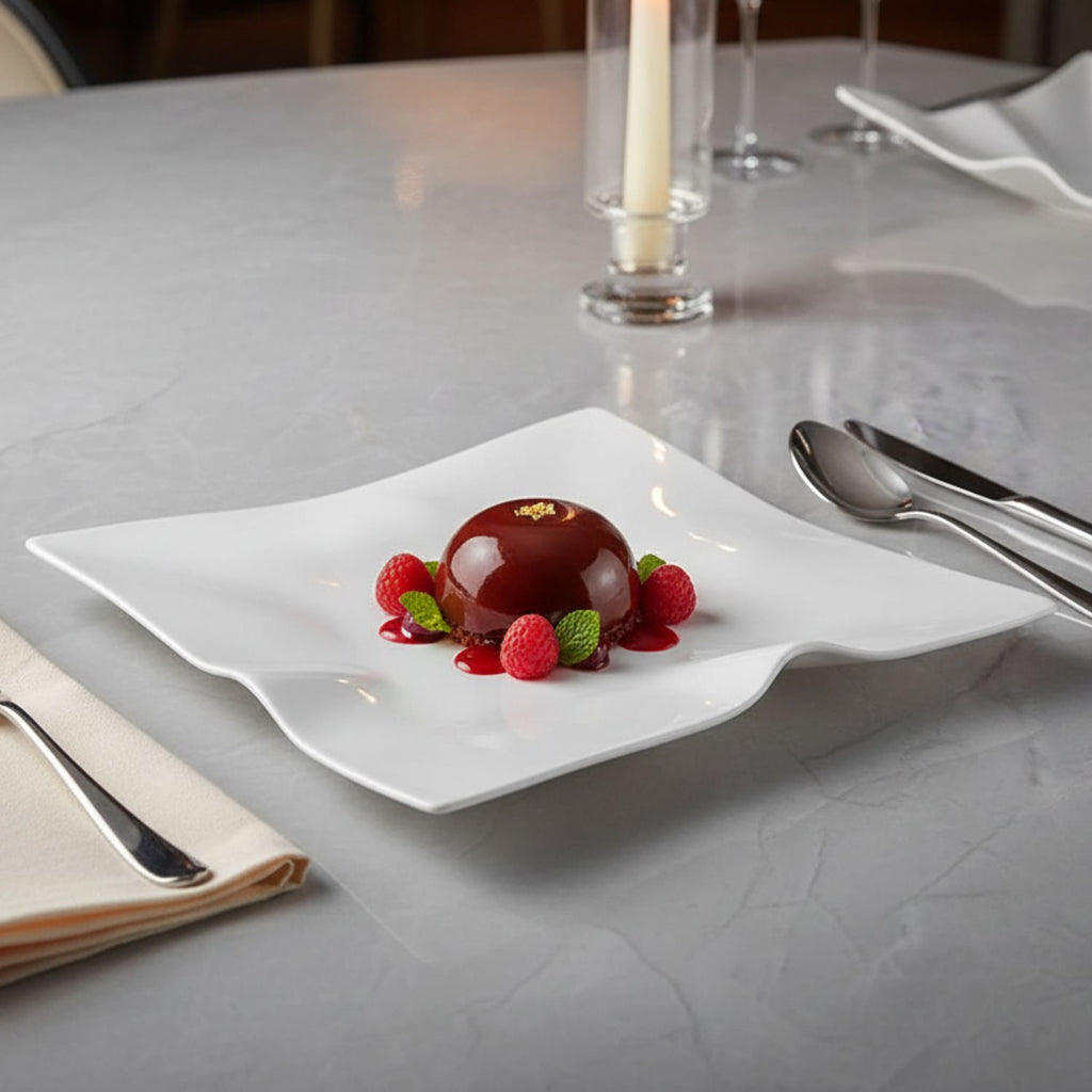A white ceramic dessert plate with a wavy, scalloped edge, styled with a chocolate mousse, raspberries, and mint on a gray marble table.