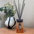 A reed diffuser bottle with an amber faceted glass body, black cap, and black reeds stands on a wooden table next to a white ceramic vase with black swirls and green eucalyptus leaves.