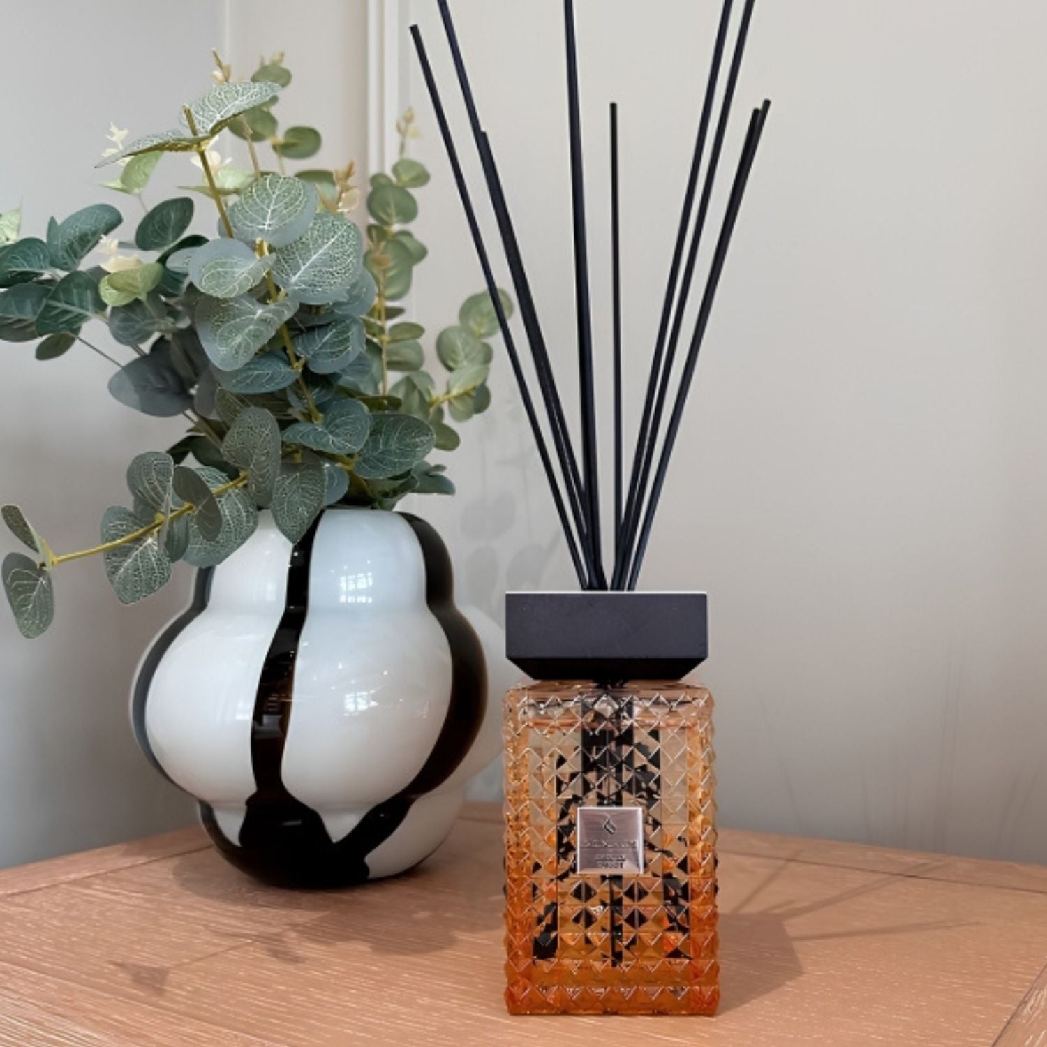 A reed diffuser bottle with an amber faceted glass body, black cap, and black reeds stands on a wooden table next to a white ceramic vase with black swirls and green eucalyptus leaves.