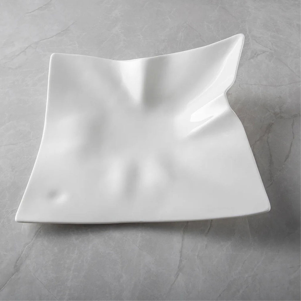 A white square ceramic serving platter with a wavy, sculptural design and a glossy finish, displayed on a gray marble-textured surface.