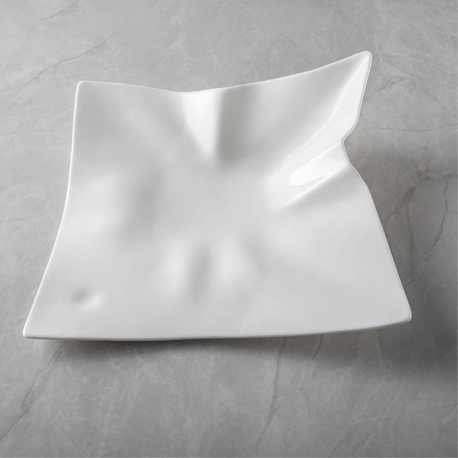 A white square ceramic serving platter with a wavy, sculptural design and a glossy finish, displayed on a gray marble-textured surface.