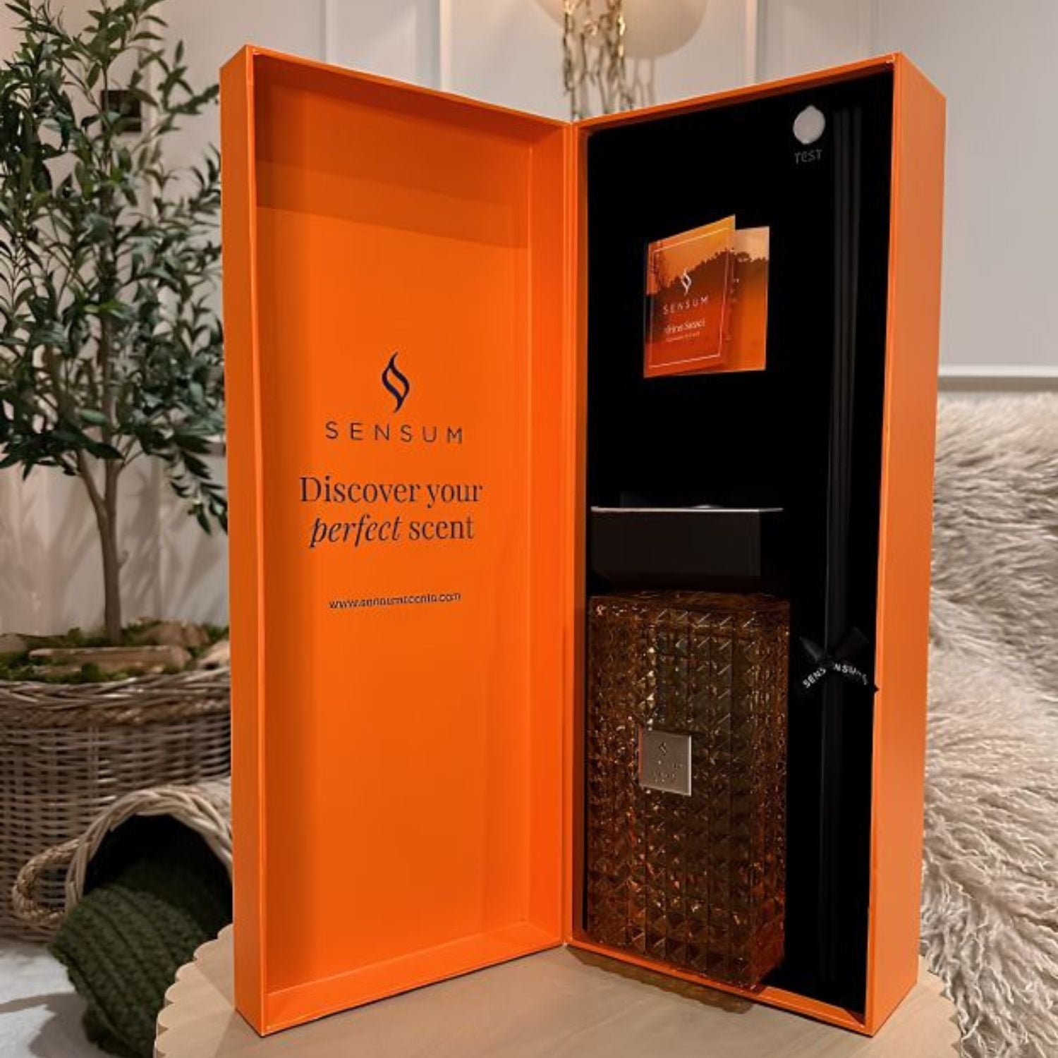An open bright orange Sensum gift box containing a textured amber-brown perfume bottle and a small card, set on a tabletop with a plant and soft furnishings in the background.