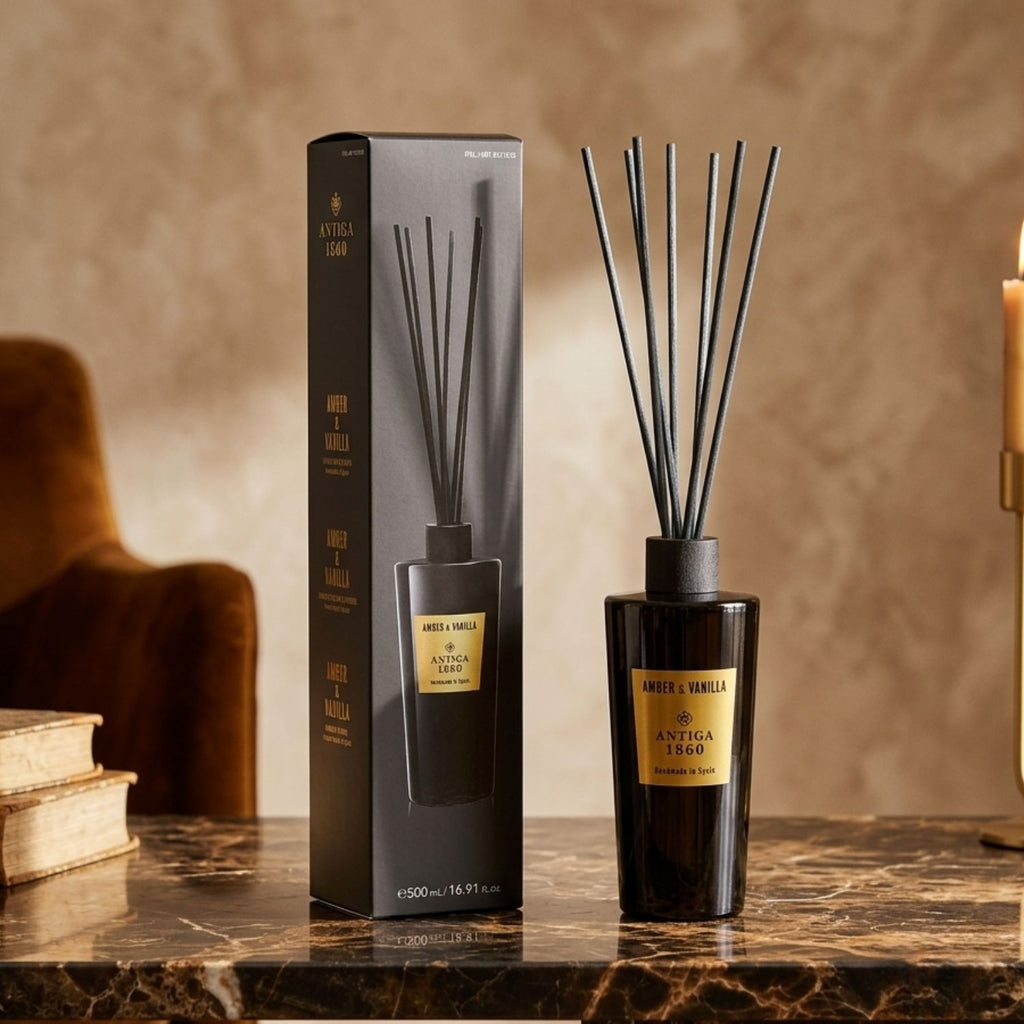 A reed diffuser bottle with black reeds stands beside its matching black box on a polished marble surface. The bottle and box are dark glass with a gold label reading 'Amber & Vanilla SYNTIGA 1950'.
