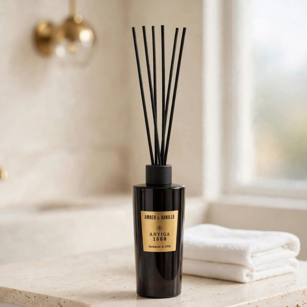 A reed diffuser bottle with black reeds stands on a light stone surface beside folded white towels, with a softly blurred bathroom background and a brass wall fixture.