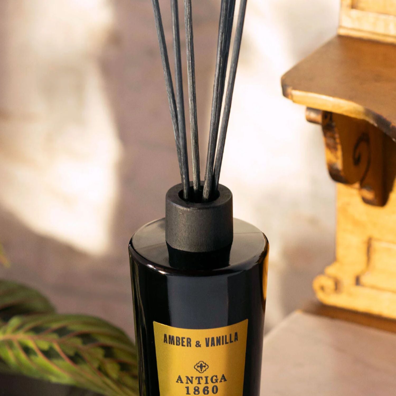 A close-up of a reed diffuser bottle with dark reeds, featuring a black glass bottle and cap with a gold label that reads 'AMBER & VANILLA' and 'ANTIGA 1860'.