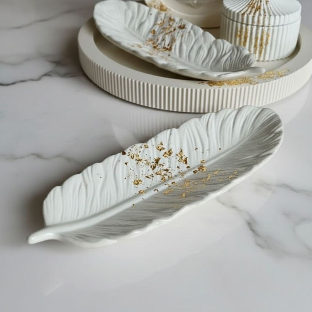 A white ceramic feather-shaped dish with gold leaf accents, displayed on a white marble surface with a matching ribbed tray and container in the background.