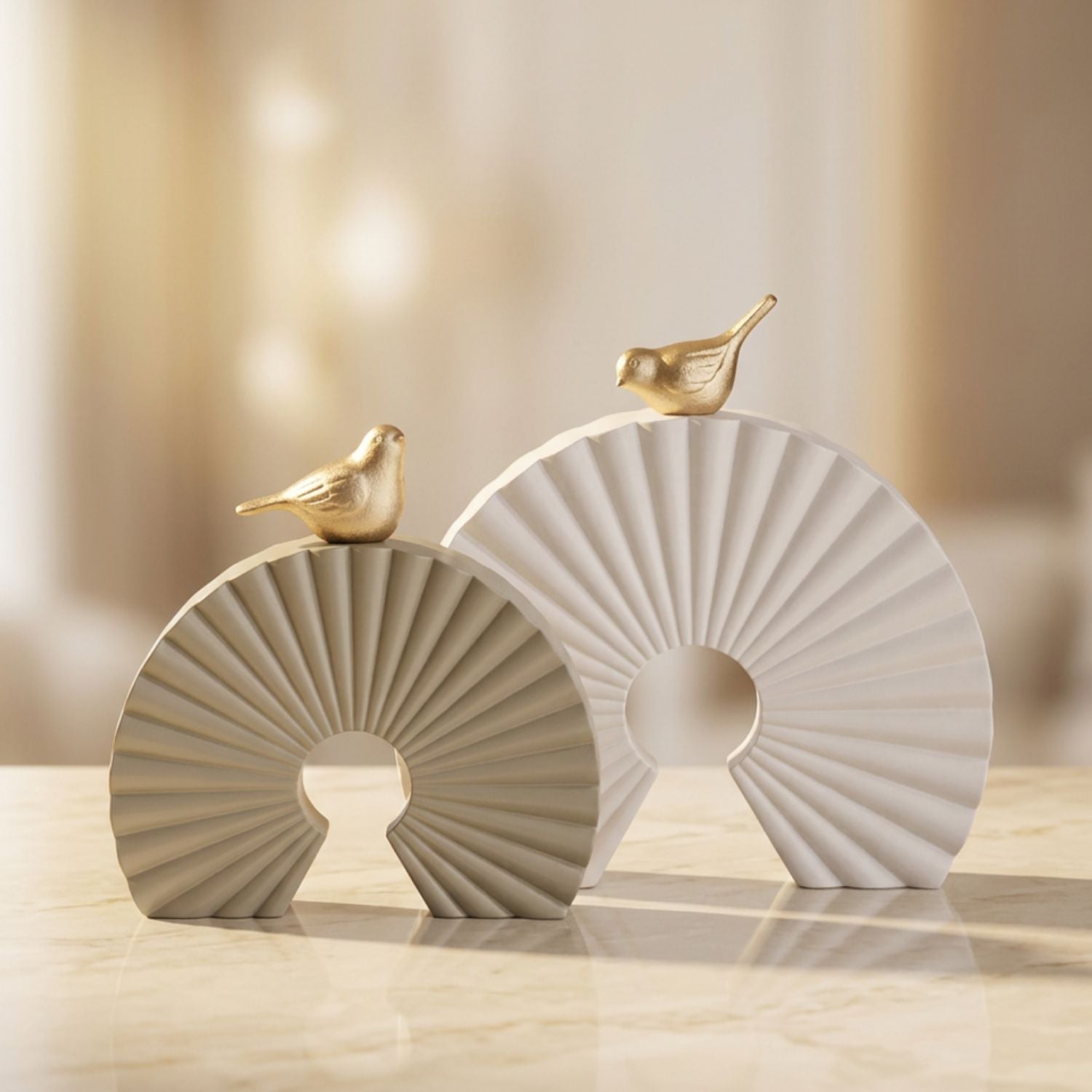 Two decorative ceramic fan-shaped sculptures in warm beige and off-white, each topped with a small metallic gold bird, displayed on a light marble surface against a softly blurred cream background.