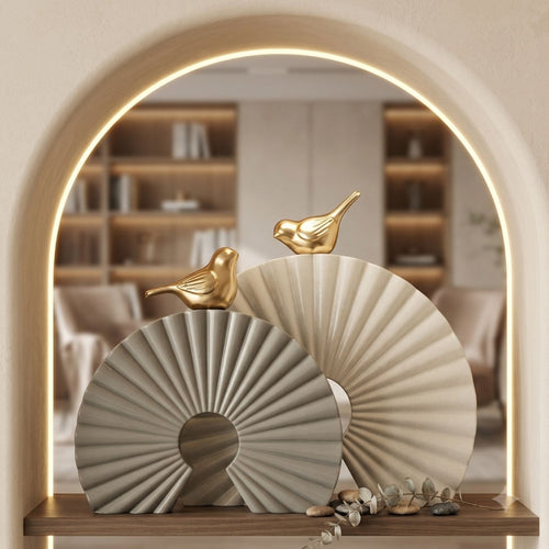Decorative fan-shaped wall art pieces in warm beige and light gray tones with small gold bird sculptures, displayed on a wooden shelf in front of an arched mirror showing a softly lit interior.