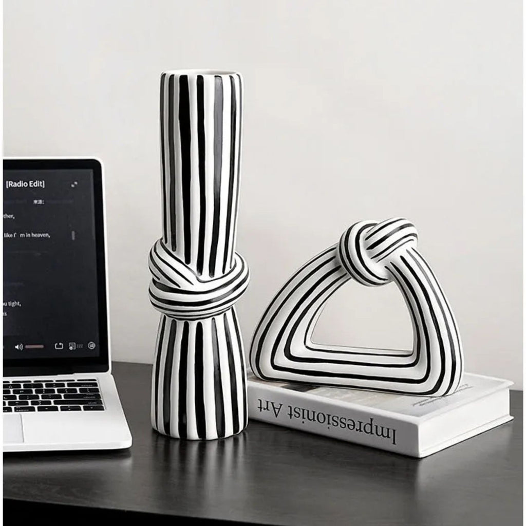 Two modern black and white striped ceramic decorative sculptures, one cylindrical with a knotted center and one triangular with a knotted top, displayed on a dark tabletop next to an open laptop and a book.