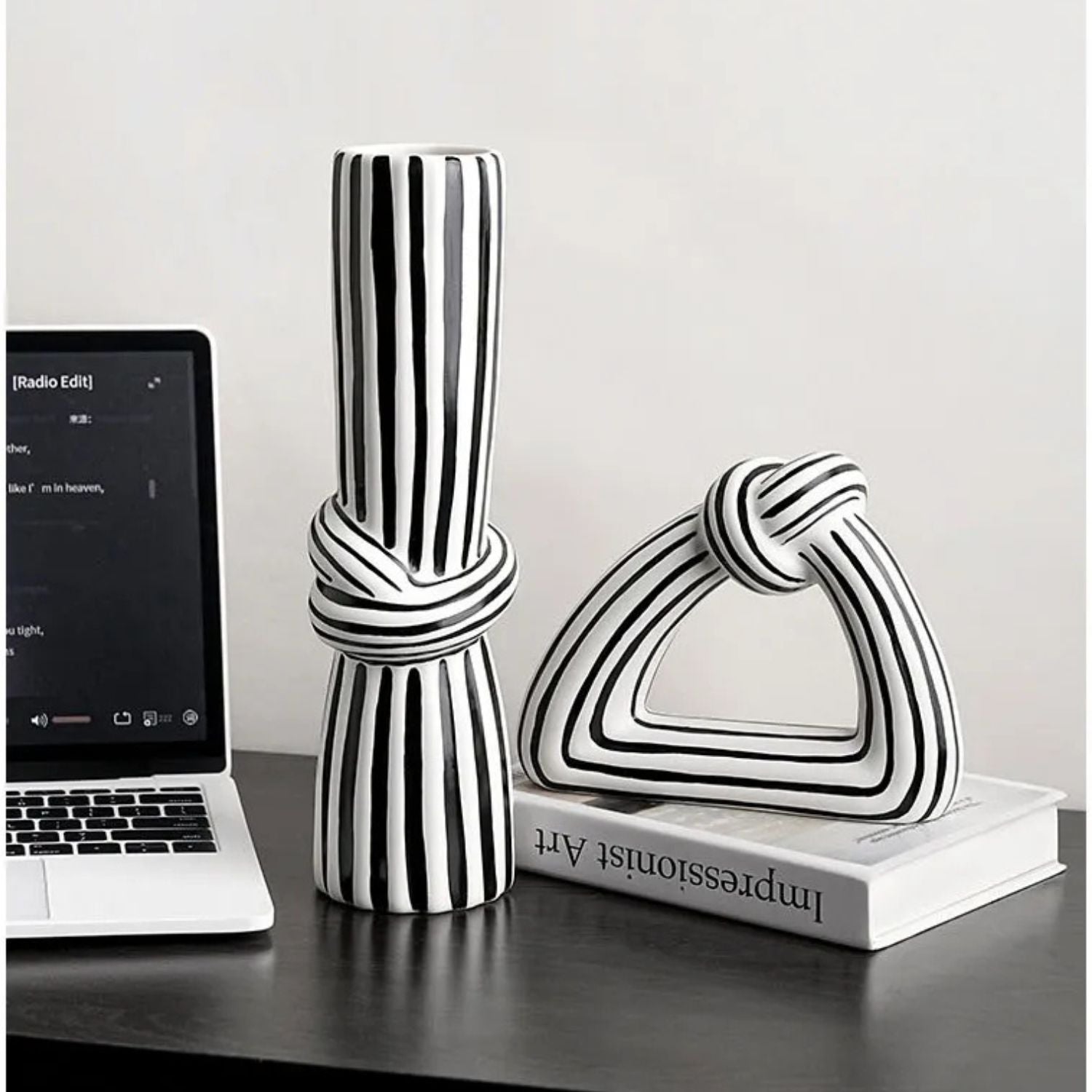 Two modern black and white striped ceramic decorative sculptures, one cylindrical with a knotted center and one triangular with a knotted top, displayed on a dark tabletop next to an open laptop and a book.