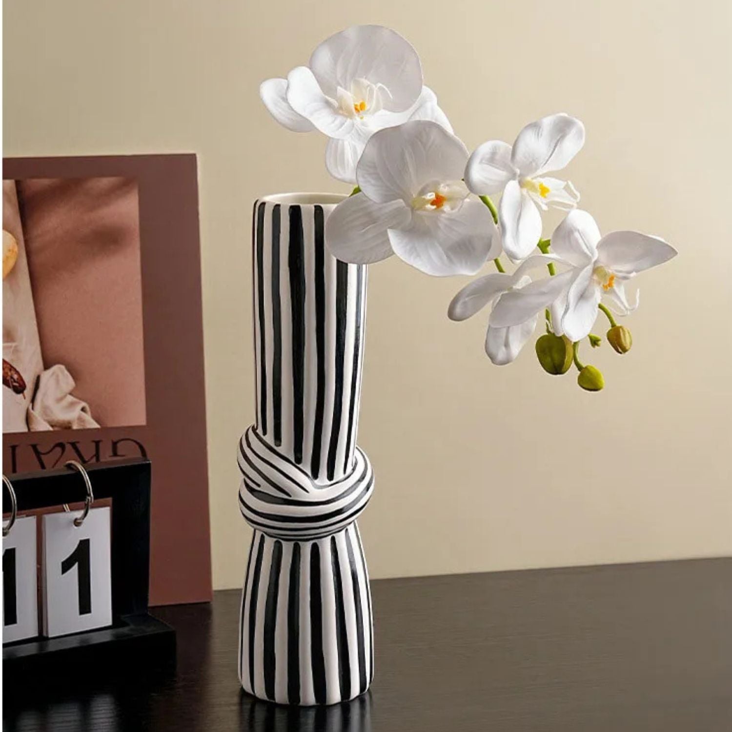 A modern black and white vertically striped ceramic vase with a knotted center detail, holding white orchid flowers, set on a dark tabletop next to a calendar and framed picture.