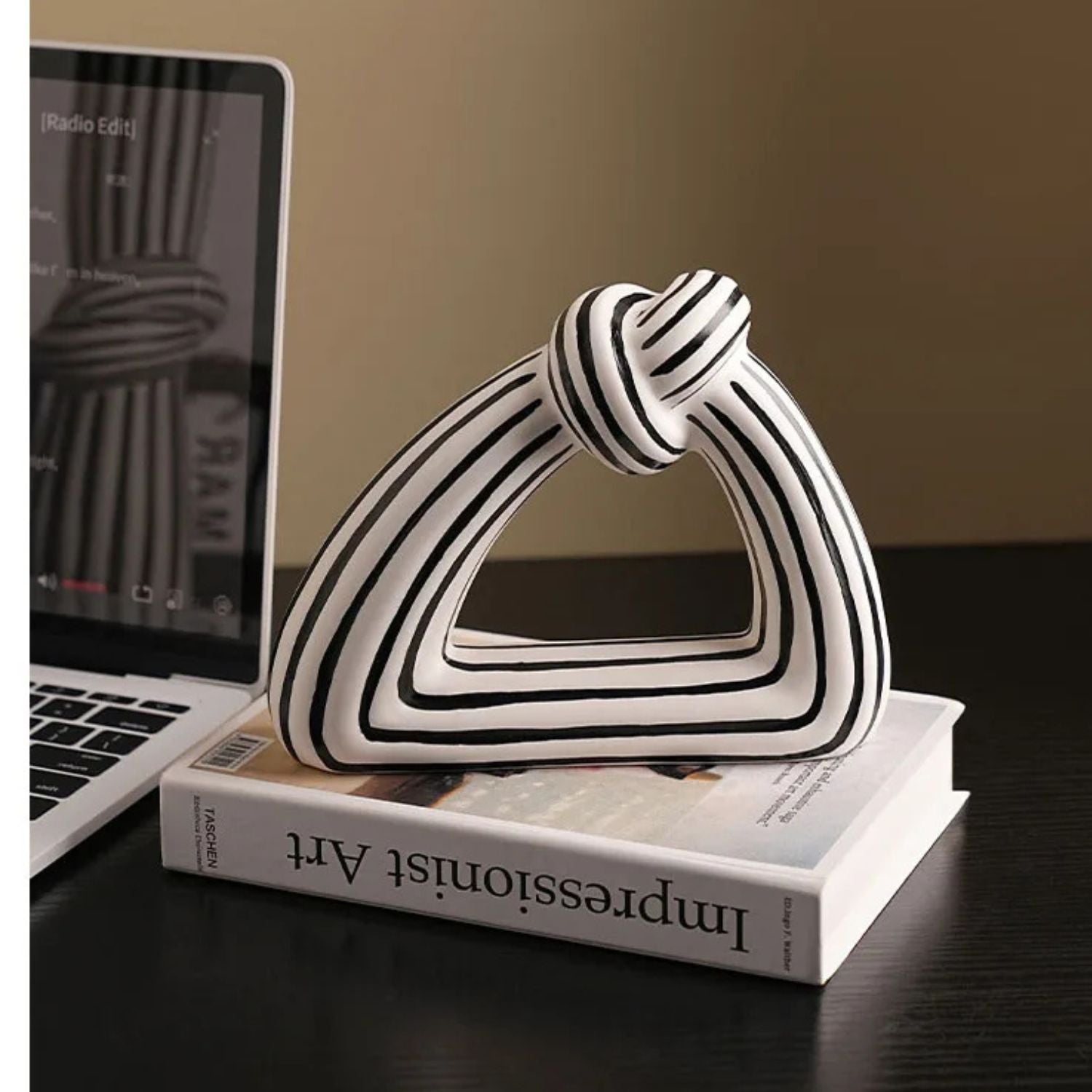 A white ceramic decorative sculpture with black concentric stripes and a knotted top detail, displayed on top of a book on a dark wooden table next to an open laptop.