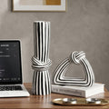A black and white striped ceramic decorative set on a wooden table, featuring a rolled scroll vase and a knot-shaped sculpture resting on a white book, styled with a laptop and gold candle holders against a textured gray wall.
