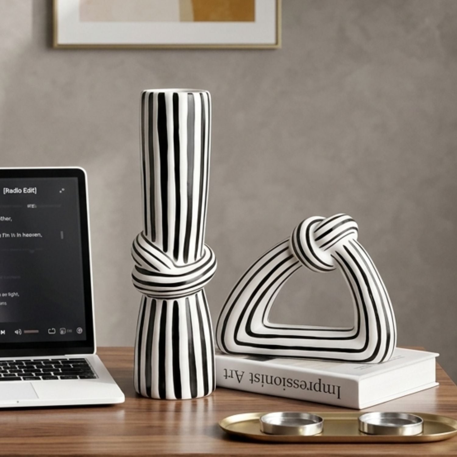 A black and white striped ceramic decorative set on a wooden table, featuring a rolled scroll vase and a knot-shaped sculpture resting on a white book, styled with a laptop and gold candle holders against a textured gray wall.