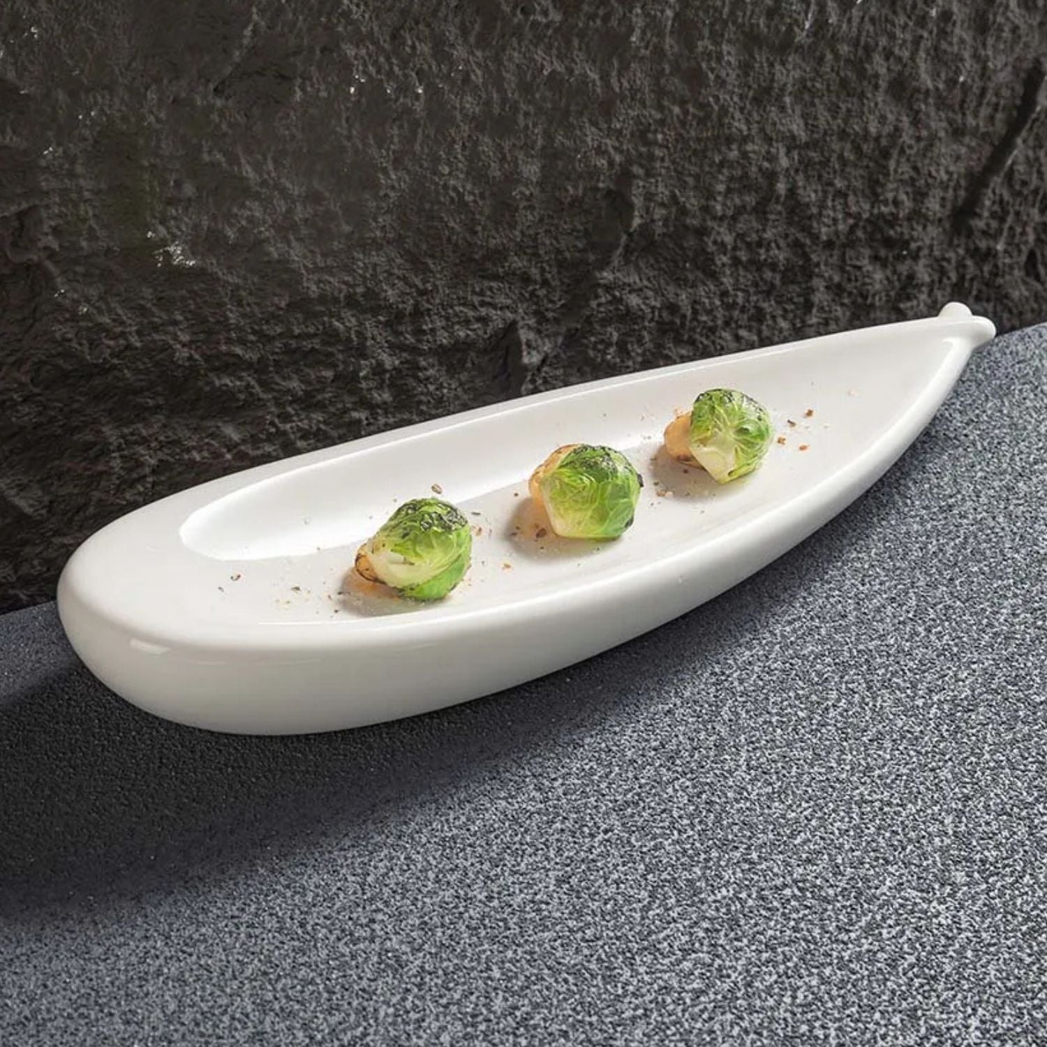 A white ceramic leaf-shaped serving platter holding three Brussels sprouts, set on a textured gray surface against a dark gray background.