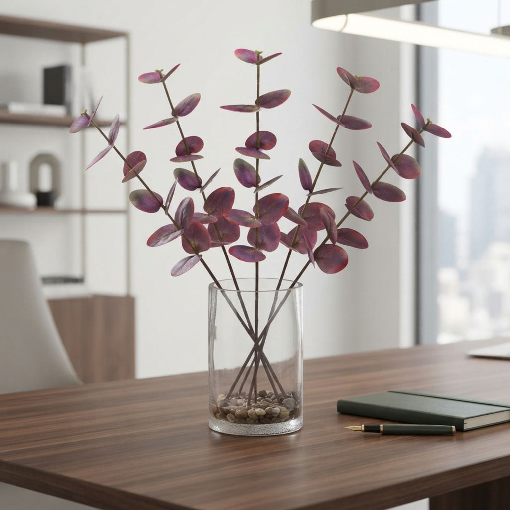 A clear cylindrical glass vase filled with pebbles and stems, holding a bouquet of pinkish-purple eucalyptus branches, placed on a wooden desk in a bright modern office setting.