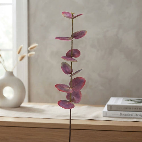 A single artificial eucalyptus stem with dusty burgundy-purple leaves stands upright on a light wood table in a softly styled interior setting.