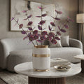 A white ceramic vase with gold trim holding purple dried branches, placed on a round white marble-top table in a modern living room setting.