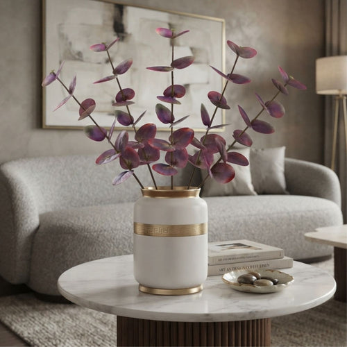 A white ceramic vase with gold trim holding purple dried branches, placed on a round white marble-top table in a modern living room setting.