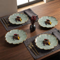 Four scalloped ceramic plates with a light green textured glaze and gold trim, arranged on a dark wood table with gray placemats and gold-toned cutlery.