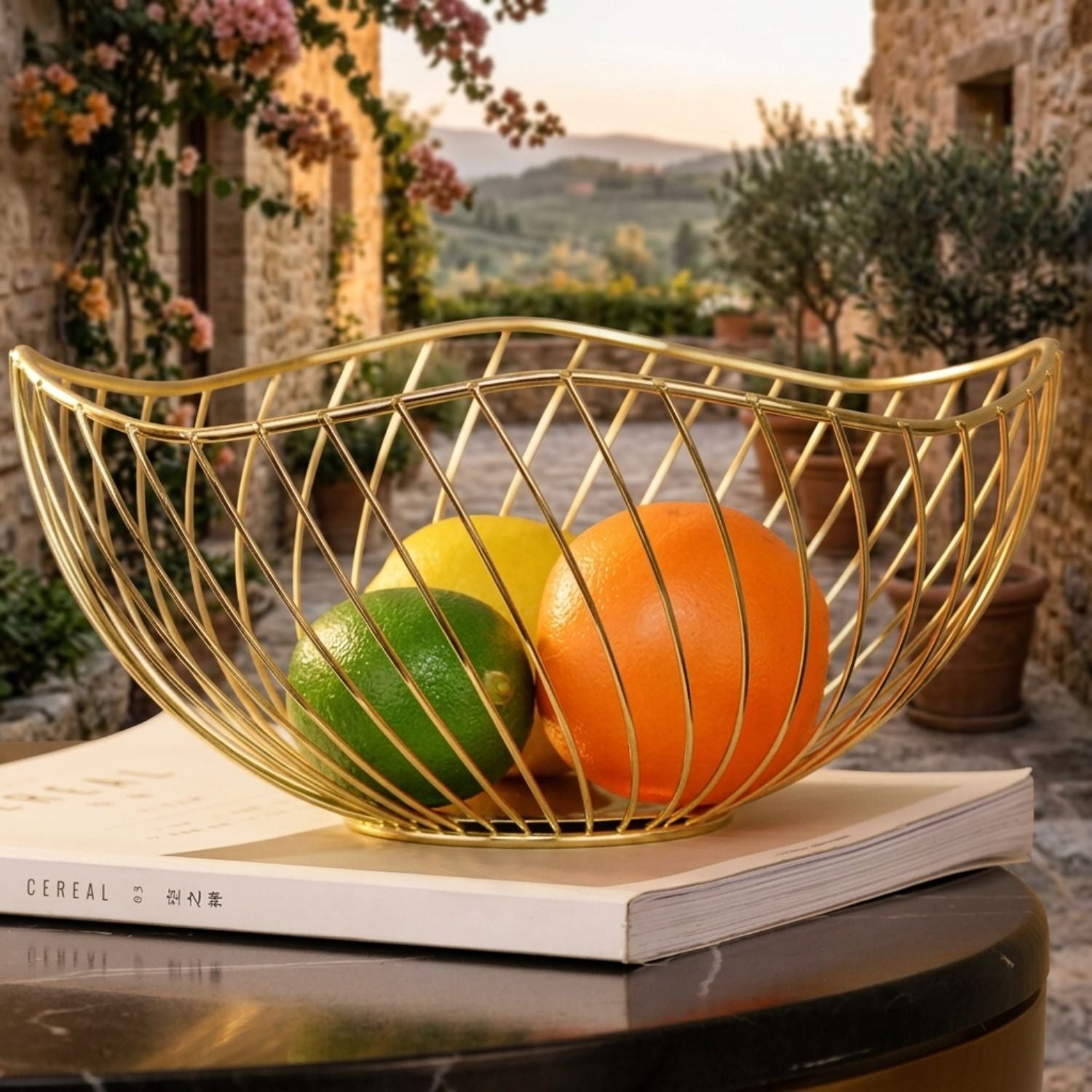 A gold wire bowl filled with citrus fruits sits on a white book atop a dark marble table, set against a blurred outdoor stone courtyard background.