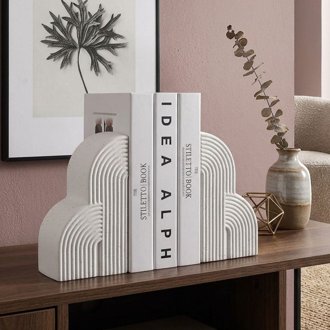 White cloud-shaped bookends holding three books on a wooden console table in a styled room with a pink wall, framed art, a vase, and a chair.